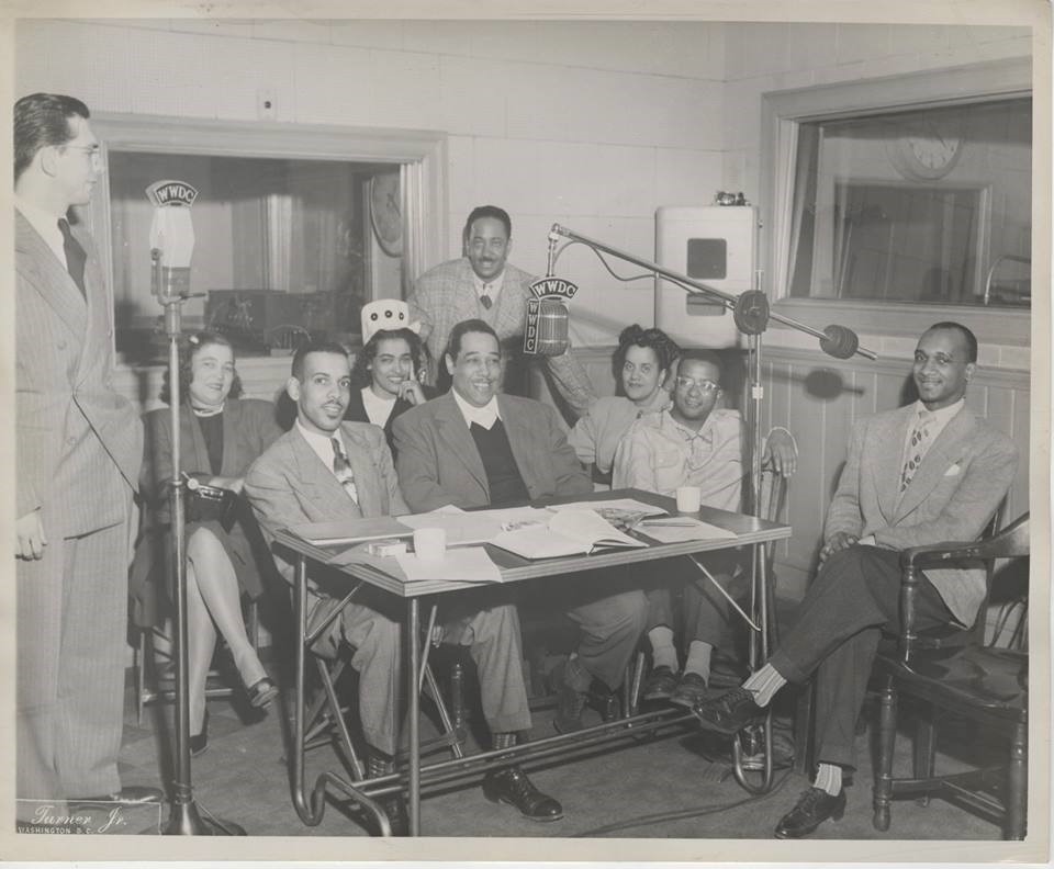 Willis Conover standing at radio microphone with seated musicians